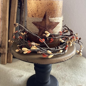 May include: A decorative candle holder with a rustic aesthetic. It features a brown candle wrapped with twine, a rusty star, and a garland of berries. The holder is on a round, wooden base with a black pedestal.