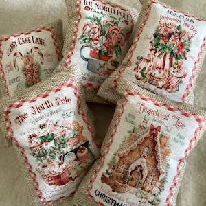 May include: A collection of burlap Christmas-themed sachets. Each sachet features a different vintage-style design with text like "Candy Cane Lane" and "Christmas Cottage." The designs include candy canes, gingerbread houses, and cupcakes.