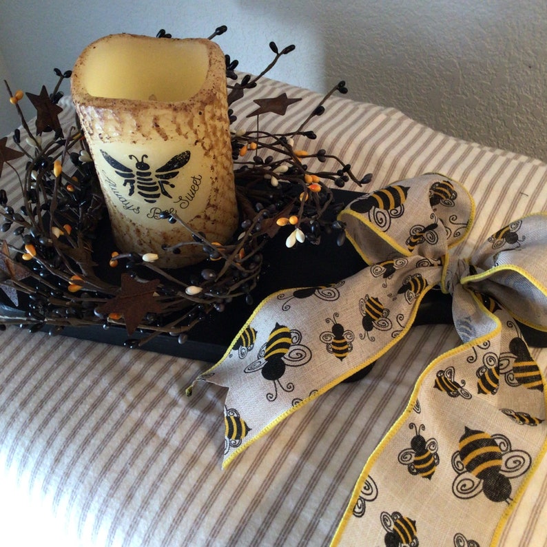 May include: A black tray with a candle in the center surrounded by a wreath of twigs and small white and orange berries. A beige ribbon with a yellow border and black bees printed on it is tied in a bow on the tray.