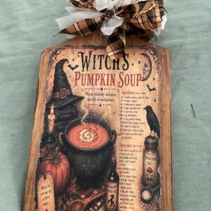 May include: A wooden cutting board featuring a printed graphic titled Witch's Pumpkin Soup, depicting a cauldron, a pumpkin, a raven, and a recipe. The board is decorated with a brown plaid and white fabric bow tied at the top handle.