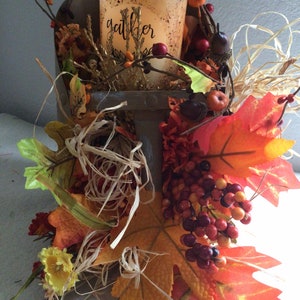 May include: A wooden box filled with fall foliage, including red berries, brown acorns, and orange and yellow leaves. A candle with the word "gather" on it sits in the box.