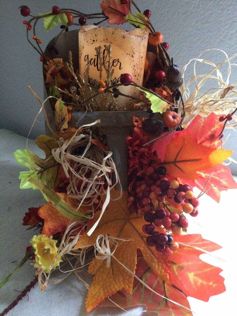 May include: A wooden box filled with fall foliage, including red berries, brown acorns, and orange and yellow leaves. A candle with the word "gather" on it sits in the box.