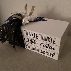 May include: A white wooden block stack featuring the black text "TWINKLE TWINKLE Little Star Do You know how Loved You are?" adorned with a decorative bow made of black ribbon with white text, gold ribbon, and white glittery star accents.
