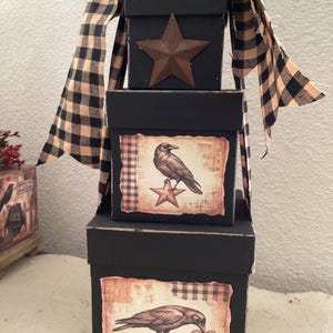 May include: A stack of three black decorative boxes, each with a different design. The top box has a rusty star, the middle box features a raven and star, and the bottom box shows a raven with a heart. A black and tan plaid ribbon is draped over the boxes.