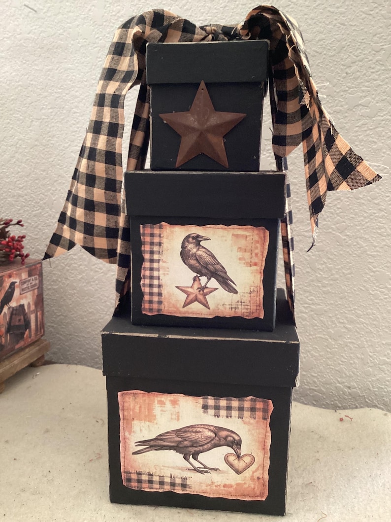 Peut inclure: Une pile de trois bo&icirc;tes carr&eacute;es noires d&eacute;cor&eacute;es d'un ruban &agrave; carreaux beige et noir. La bo&icirc;te sup&eacute;rieure pr&eacute;sente une &eacute;toile en m&eacute;tal, celle du milieu montre un corbeau sur une &eacute;toile, et la bo&icirc;te inf&eacute;rieure illustre un corbeau tenant un c&oelig;ur.