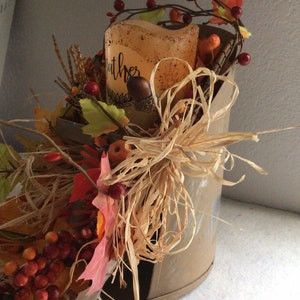 May include: A rustic metal container filled with fall decor, including faux berries, leaves, and a candle with the word "gather" written on it. The container is decorated with a brown, straw-like material.
