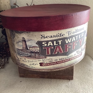 May include: A round, vintage-style container with a dark red lid and a cylindrical body. The container features a label with the text "Seaside Tradition SALT WATER TAFFY" and a lighthouse illustration. The words "Made Fresh on the Shore!" are also visible.
