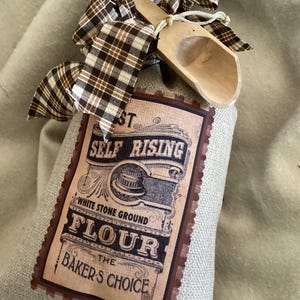 May include: A burlap bag of flour with a vintage label that reads "Self Rising Flour, White Stone Ground, The Baker's Choice." A small wooden scoop and a brown plaid bow are tied to the top of the bag. The bag is sitting on a light beige surface.
