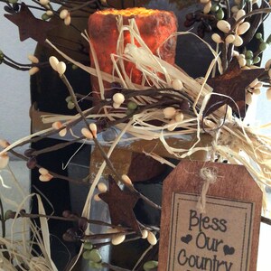 May include: A rustic wooden sign with the text "Bless Our Country Home" in black lettering. The sign is decorated with a brown burlap bow and is surrounded by a wreath of twigs, faux berries, and small white flowers.