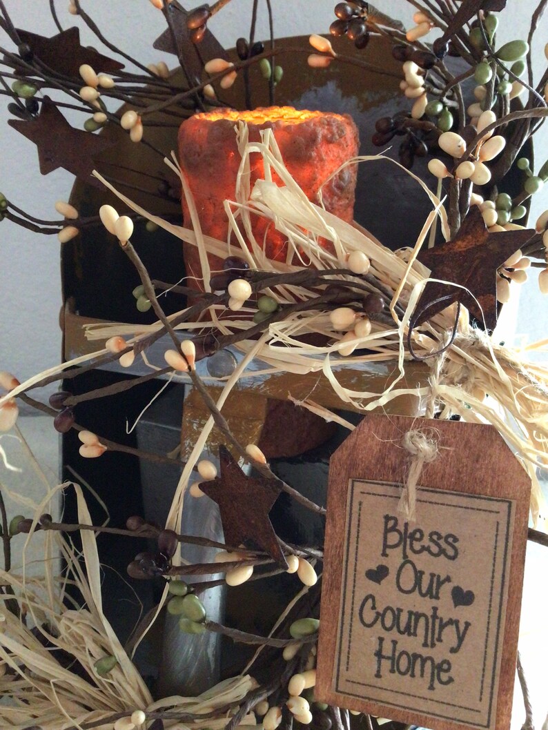 May include: A rustic wooden sign with the text "Bless Our Country Home" in black lettering. The sign is decorated with a brown burlap bow and is surrounded by a wreath of twigs, faux berries, and small white flowers.