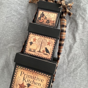 Peut inclure: Une pile de trois bo&icirc;tes en bois noir attach&eacute;es avec un ruban &agrave; carreaux beige et noir effiloch&eacute;. Chaque bo&icirc;te pr&eacute;sente une &eacute;tiquette d&eacute;corative avec des illustrations de moutons, de corbeaux, d'&eacute;toiles et le texte "Blessed Beyond Measure" et "Primitive Kitchen".