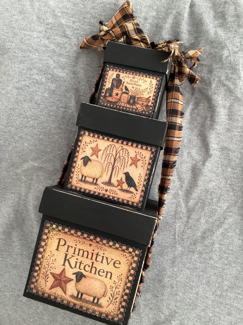 Peut inclure: Une pile de trois bo&icirc;tes en bois noir attach&eacute;es avec un ruban &agrave; carreaux beige et noir effiloch&eacute;. Chaque bo&icirc;te pr&eacute;sente une &eacute;tiquette d&eacute;corative avec des illustrations de moutons, de corbeaux, d'&eacute;toiles et le texte "Blessed Beyond Measure" et "Primitive Kitchen".