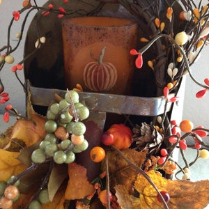 May include: A rustic metal scoop with a fall-themed arrangement. The scoop holds a brown candle with a pumpkin design. The arrangement includes fall leaves, berries, and twigs.
