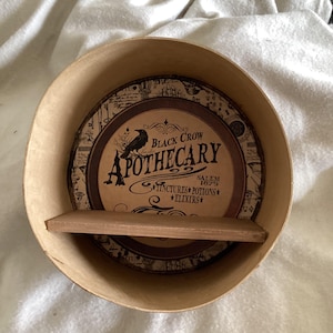 May include: A round, light brown apothecary shelf with a circular, decorative interior. The interior features a label with the text "Black Crow Apothecary" and a crow illustration. The shelf includes a small, rectangular shelf.