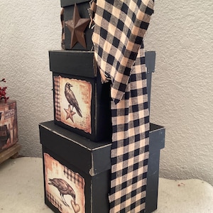 Peut inclure: Une pile de trois bo&icirc;tes carr&eacute;es noires d&eacute;cor&eacute;es d'une longue bande de tissu &agrave; carreaux vichy noir et beige. La bo&icirc;te sup&eacute;rieure pr&eacute;sente une &eacute;toile en m&eacute;tal, tandis que les bo&icirc;tes du milieu et du bas affichent des &eacute;tiquettes avec des corbeaux.