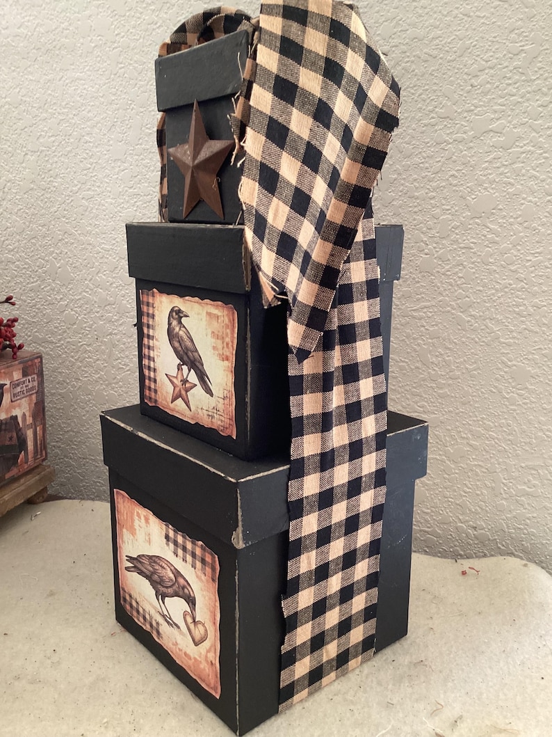 Peut inclure: Une pile de trois bo&icirc;tes carr&eacute;es noires d&eacute;cor&eacute;es d'une longue bande de tissu &agrave; carreaux vichy noir et beige. La bo&icirc;te sup&eacute;rieure pr&eacute;sente une &eacute;toile en m&eacute;tal, tandis que les bo&icirc;tes du milieu et du bas affichent des &eacute;tiquettes avec des corbeaux.