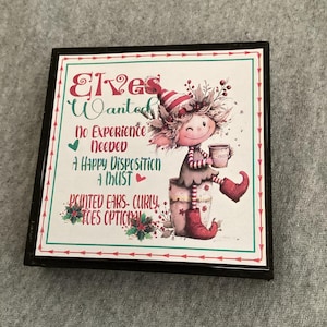 May include: Square decorative sign with a black frame. The sign features a whimsical illustration of an elf holding a mug. Text on the sign reads "Elves Wanted" with additional text about requirements. The color palette includes red, green, and white.