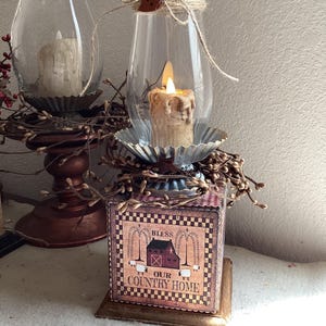 May include: A square wooden block base with a printed image of a barn, sheep, and the text BLESS OUR COUNTRY HOME. It features a metal fluted candle holder, a glass hurricane shade, a battery-operated candle, and a brown heart-shaped button.