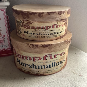 May include: Two round, vintage-style boxes stacked on top of each other. The boxes are tan with brown edges and feature the text "Campfire Marshmallows" in a vintage font. The top box has a deer graphic.