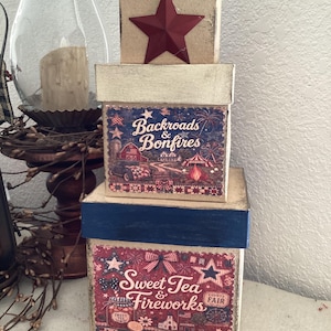 May include: A stack of three decorative boxes with patriotic themes. The top box is red with a red star. The middle box has a blue and red design with the words "Backroads & Bonfires." The bottom box has a red and blue design with the words "Sweet Tea & Fireworks."