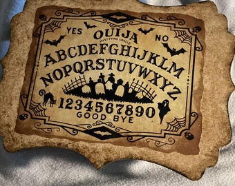 May include: A rectangular Ouija board with scalloped edges, featuring a tan, aged-paper appearance. The board displays the alphabet, numbers 0-9, the words YES, NO, and GOOD BYE, along with black illustrations of bats, spiderwebs, skulls, a cat, a ghost, and a graveyard.