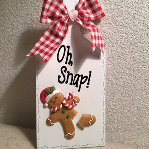 May include: White wooden sign with a red and white gingham bow. The sign says "Oh, Snap!" and features a gingerbread man with a broken leg.