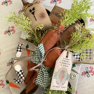 May include: A rustic Easter-themed arrangement featuring a fabric bunny, faux carrots, and greenery nestled in a muffin tin. The carrots are wrapped in green gingham fabric and tied with twine. A tag reads "Farm Fresh Carrots 4 Sale".