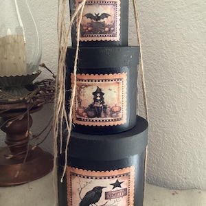 May include: A stack of three cylindrical black boxes with lids, tied together with natural jute twine. Each box features a square label with scalloped edges depicting Halloween imagery: a bat, a witch, and a crow with the text "PRIMITIVE".