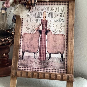 May include: A wooden framed sign featuring a print of a person standing between two sheep. The text reads: SEEKS WOOL AND FLAX AND WORKS WILLINGLY WITH HER HANDS PROVERBS 31:13. A decorative bow made of plaid and white fabric is attached to the top left corner.