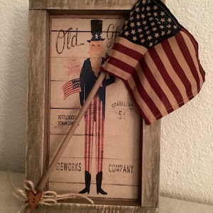 May include: A framed patriotic print featuring an illustration of Uncle Sam holding a small American flag. The print has a rustic, weathered look, with the text "Old Glory" and "Fireworks Company". A small American flag is attached to the frame.