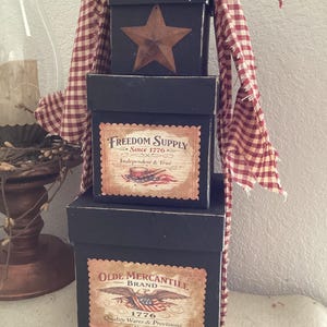 May include: Three stacked black boxes with vintage-style labels and a rusty metal star on top. The labels read "Freedom Supply Since 1776" and "Olde Mercantile Brand 1776". A red and white checkered fabric is draped over the boxes.