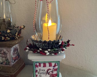 Könnte beinhalten: Ein dekorativer Weihnachts-Kerzenhalter mit einer Glas-Windlichtlaterne. Der weiße Sockel zeigt ein festliches Bild mit dem Text "Elves Wanted No Experience Needed". Die Laterne ist mit rot-weißem Garn und einem roten Pompon verziert.