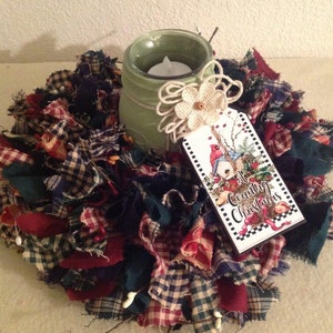 May include: A green painted mason jar candle holder with a burlap bow and a white flower. The candle holder is surrounded by a wreath made of red, green and blue plaid fabric scraps. A tag with the text "A Country Christmas" is attached to the jar.