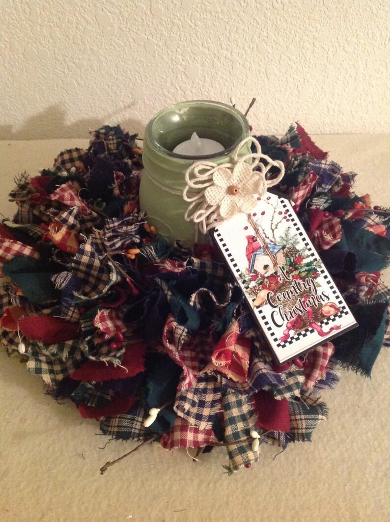 May include: A green painted mason jar candle holder with a burlap bow and a white flower. The candle holder is surrounded by a wreath made of red, green and blue plaid fabric scraps. A tag with the text "A Country Christmas" is attached to the jar.