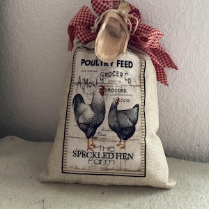 May include: A burlap sack labeled "Poultry Feed" with a vintage design featuring two chickens. The sack is tied with a red and white checkered ribbon and a small wooden scoop. The text on the sack reads "A Moll Grocer Co" and "The Speckled Hen Farm."