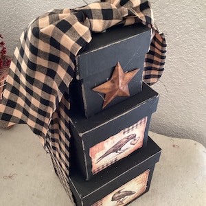 Peut inclure: Une pile de trois bo&icirc;tes carr&eacute;es noires aux bords us&eacute;s. La bo&icirc;te sup&eacute;rieure pr&eacute;sente une &eacute;toile en m&eacute;tal rouill&eacute;. Les bo&icirc;tes du milieu et du bas affichent des &eacute;tiquettes rectangulaires avec des illustrations de corbeaux noirs. Un ruban en tissu &agrave; carreaux beige et noir.