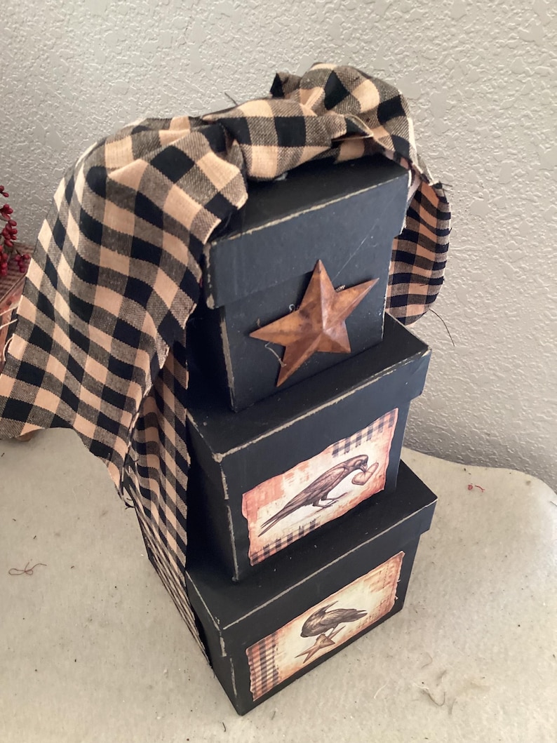 Peut inclure: Une pile de trois bo&icirc;tes carr&eacute;es noires aux bords us&eacute;s. La bo&icirc;te sup&eacute;rieure pr&eacute;sente une &eacute;toile en m&eacute;tal rouill&eacute;. Les bo&icirc;tes du milieu et du bas affichent des &eacute;tiquettes rectangulaires avec des illustrations de corbeaux noirs. Un ruban en tissu &agrave; carreaux beige et noir.