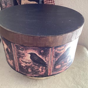 May include: A round, black-topped decorative box with a dark wood-tone band. The box's sides feature a vintage-style print with a raven, skulls, and other gothic imagery. The box has a rustic, antique appearance.