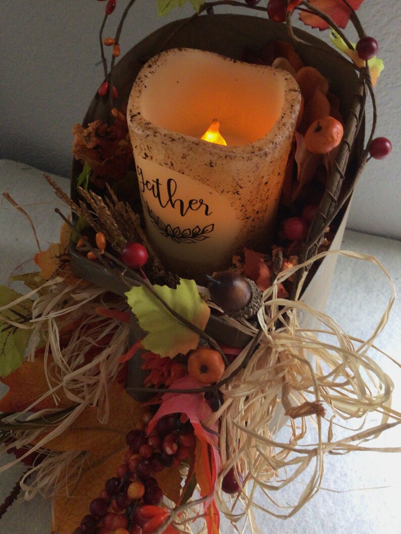 May include: A brown metal bucket filled with fall-themed decorations, including faux leaves, berries, and a lit candle. The candle is a pillar candle with a white, textured surface and the word "gather" written on it.