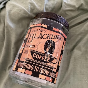 May include: A clear glass jar with a dark, textured lid. The label features a black bird illustration, the text CRANKY BLACKBIRD BRAND DARK ROAST COFFEE, and the phrase SOMETHING TO GROW ABOUT, all set against a rectangular orange and black border.