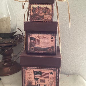 May include: A stack of three square dark brown boxes tied with tan raffia ribbon. Each box features a printed label with patriotic imagery, including American flags, stars, and text such as Celebrate Freedom, America the Beautiful, and Land That I Love.