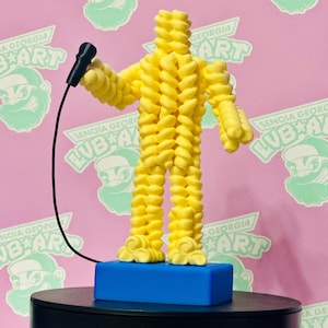 May include: A yellow sculpture of a person holding a microphone, made from twisted pasta. The figure stands on a blue base atop a black surface. The background is pink with a repeating logo that says "SENOIA GEORGIA LUB ART".