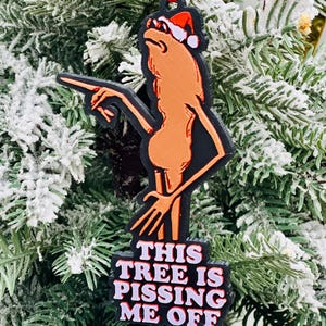 May include: A Christmas tree ornament featuring a cartoon figure with a Santa hat, pointing. The ornament is black and orange with the text "THIS TREE IS PISSING ME OFF" in white and pink.