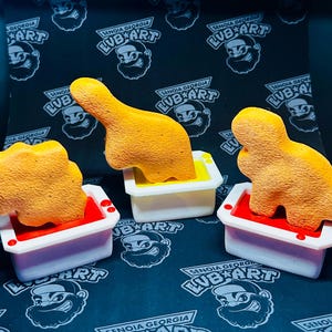 Puede incluir: Tres juguetes de dinosaurio de color naranja están sumergidos en bañeras blancas llenas de líquidos rojo, amarillo y naranja. Las bañeras están sobre un fondo negro con un patrón repetido de un rostro de hombre con barba y sombrero.