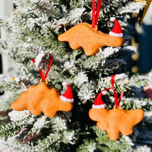 Könnte beinhalten: Drei orangefarbene Dinosaurier-Weihnachtsornamente mit roten Bändern und kleinen Weihnachtsmützen. Die Ornamente hängen an einem schneebedeckten Weihnachtsbaum und schaffen eine festliche Urlaubsszene.