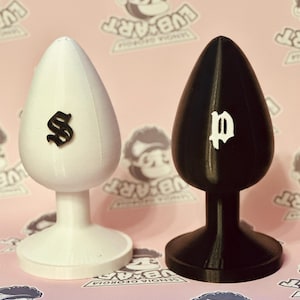 Puede incluir: Dos tapones anales, uno blanco con una "S" negra y otro negro con una "p" blanca. Los tapones tienen forma de lágrima y una base acampanada. El fondo es rosa con texto repetido "LUB ART" y "SENOIA GEORGIA".