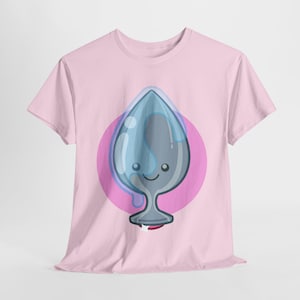 May include: A light pink t-shirt featuring a cartoon illustration of a gray and blue teardrop-shaped object with a smiling face, set against a pink circle. The design has a playful, whimsical style.