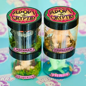 May include: Two clear jars with black lids, each containing a miniature cryptid figure. One jar features a Mothman figure, the other a Chupacabra. The jars are labeled "Adopt-A-Cryptid" and include the names of the cryptids.