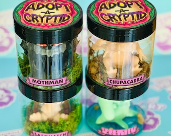 May include: Two clear jars with black lids, each containing a miniature cryptid figure. One jar features a Mothman figure, the other a Chupacabra. The jars are labeled "Adopt-A-Cryptid" and include the names of the cryptids.