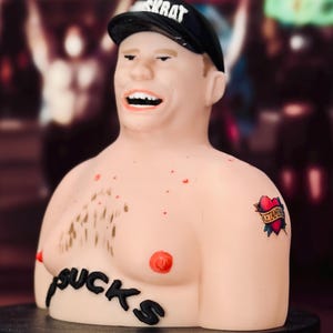 May include: A light-skinned male torso figurine wearing a black cap with the text MUSKRAT. The figure has red nipples, chest hair, and the word SUCKS written across the chest. A heart tattoo on the left shoulder contains the text KETAMINE.
